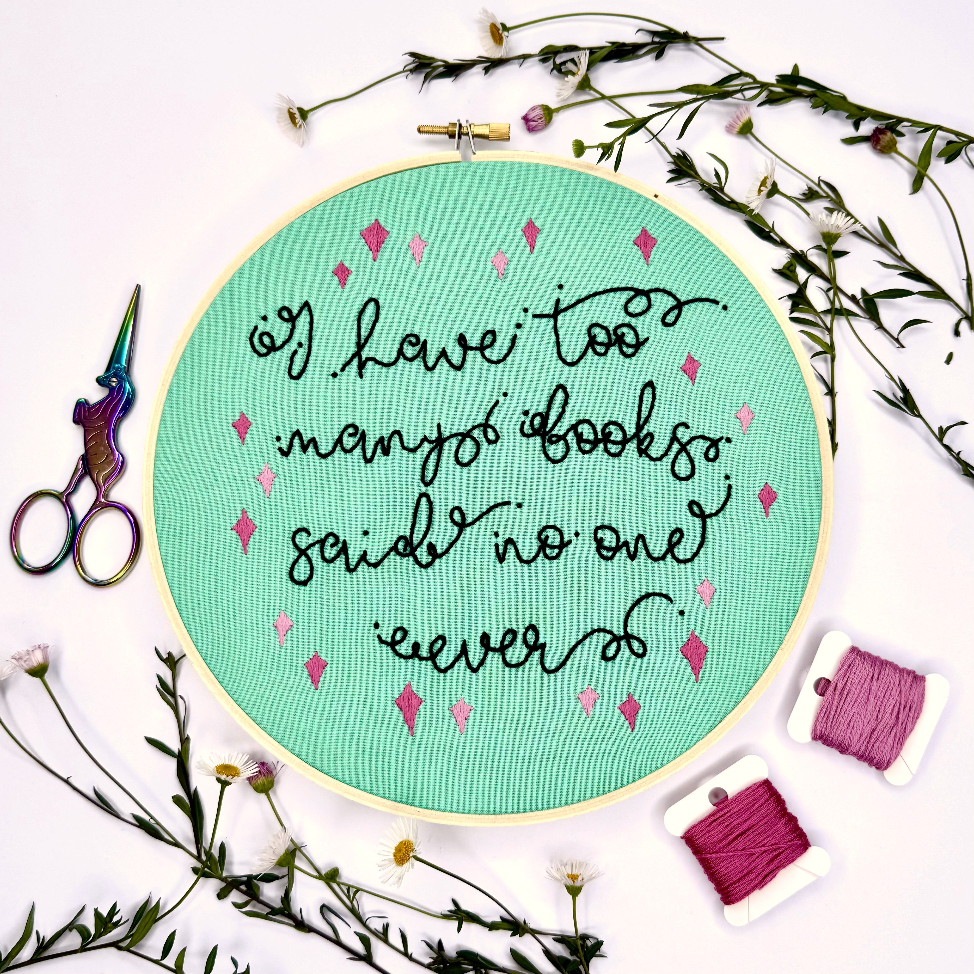 Embroidery hoop with text 'I have too many books, said no one ever.' surrounded by flowers and sewing supplies.