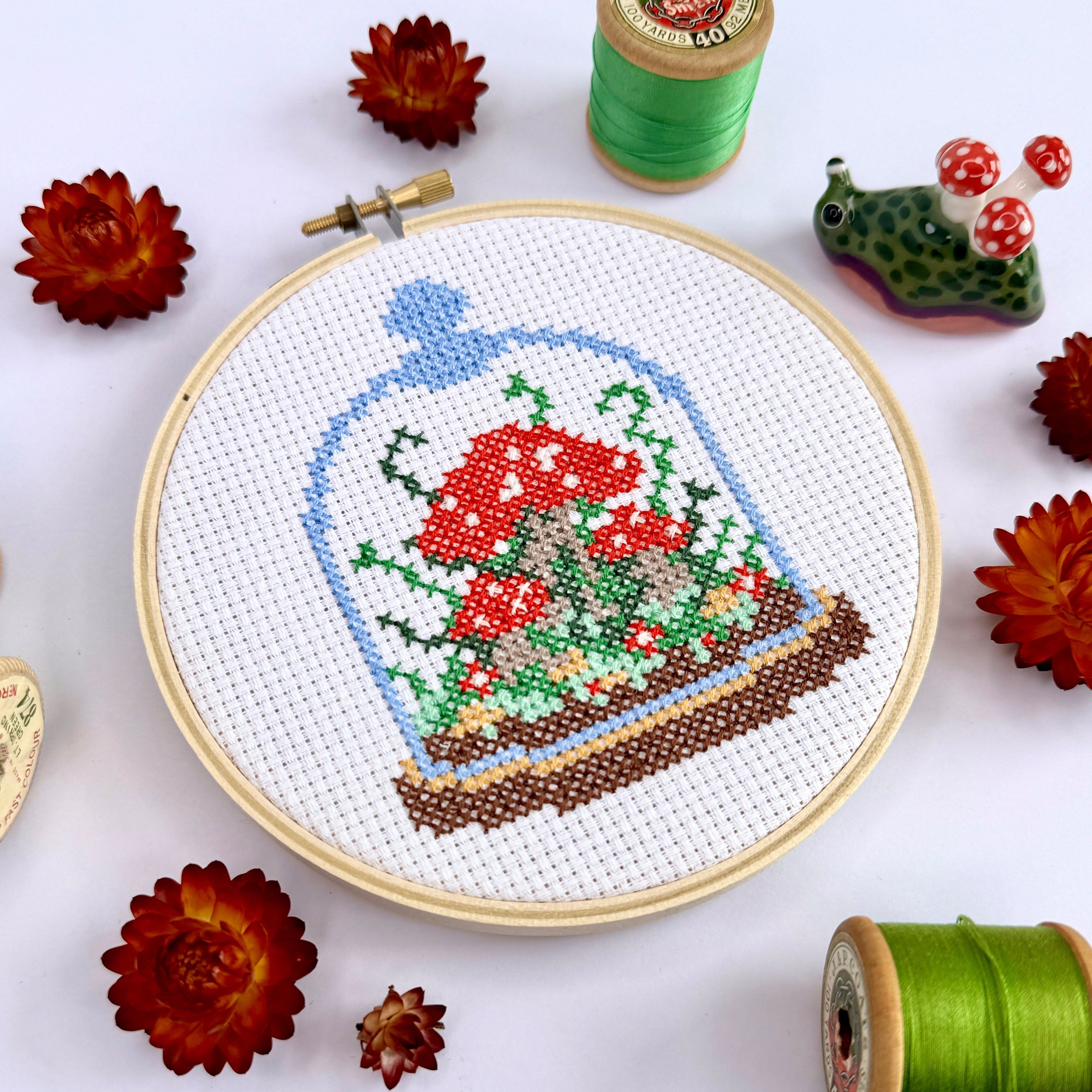 Cross-stitched design in an embroidery hoop of mushrooms and plants in a glass dome on a white background with spools of thread and dried flowers.