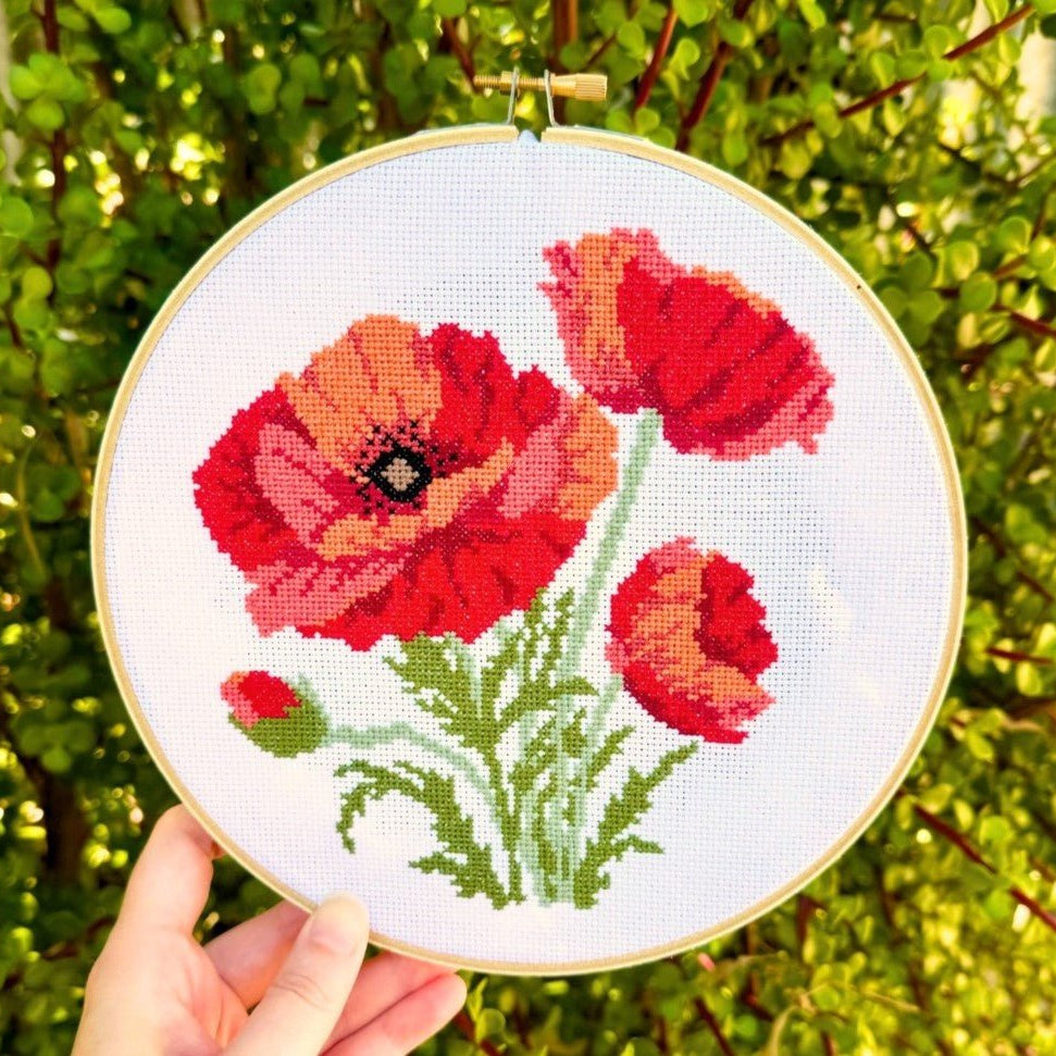 Cross stitch kit depicting a design of red poppies on a white fabric background, held up against a blurred greenery background.