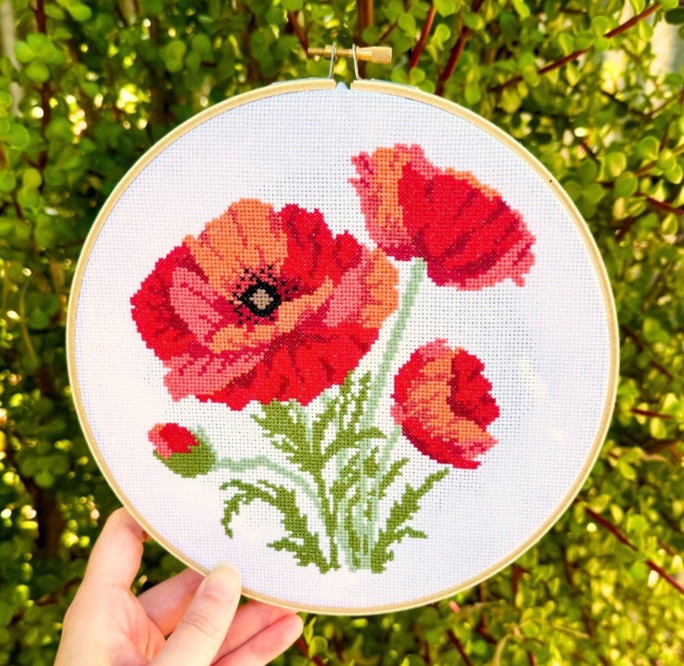 Cross stitch kit depicting a design of red poppies on a white fabric background, held up against a blurred greenery background.