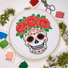 A circular embroidery hoop displays a completed cross-stitch design of a sugar skull with a pink rose crown, surrounded by spools of thread, scissors, and small green leaves on a white background.